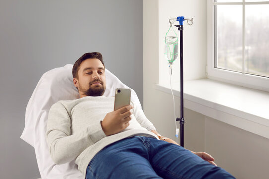 Happy relaxed young man looking at his mobile phone and watching a video while lying on a medical couch in the hospital and receiving medication through an intravenous drip