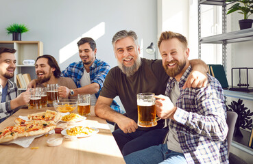 Happy male friends drinking beer at home together. Group of smiling men celebrating meeting, drinking beer and talking to each other while sitting at table at home and looking at camera