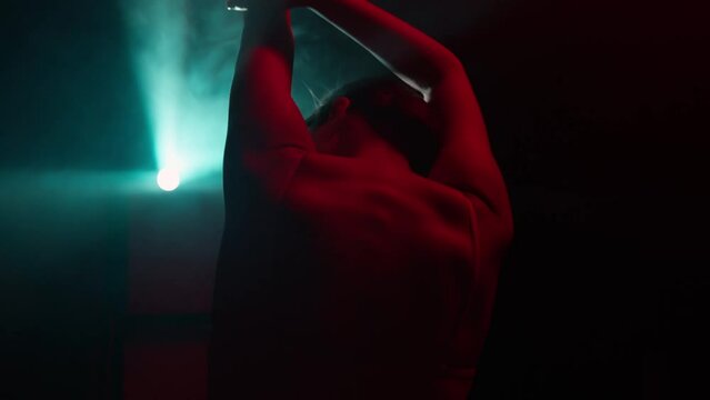 Female dancer sensually moves, dancing in dark in light rays, gesturing hands touching herself. Back view of graceful woman performing elements of modern choreography in neon illumination