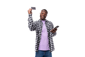positive smiling young african man with short haircut in plaid shirt holding card mockup and phone...