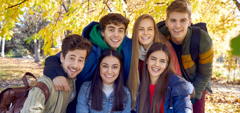 Group portrait of cheerful friends in a golden autumn park. Six happy joyful young people standing under the branches with yellow leaves, all together looking at the camera and smiling