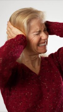 Vertical video of unhappy displeased mature older woman covering her ears with her hands because of loud noise standing model isolated on white background in studio