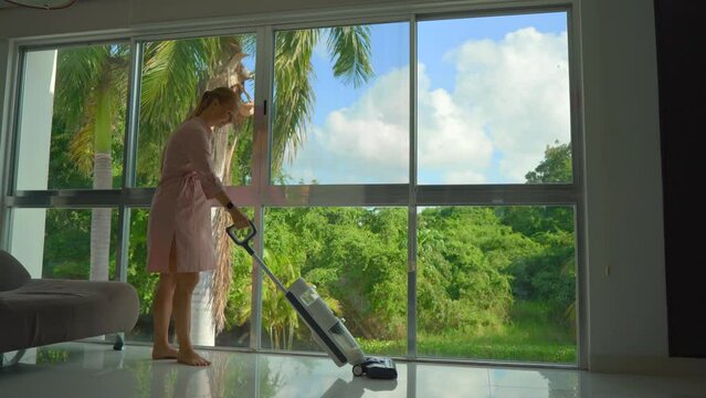 A stock video showcases a pregnant woman wearing a pink dress as she gracefully cleans the floor using a cordless electric mop vacuum cleaner. The camera captures her standing by a panoramic window