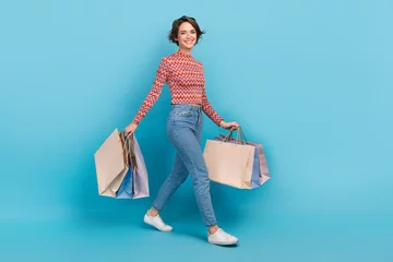 Gordijnen Full length photo of excited shiny lady wear striped top walking holding shoppers isolated blue color background © deagreez