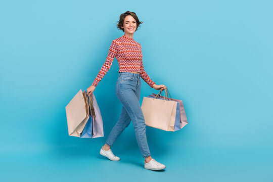 Full length photo of excited shiny lady wear striped top walking holding shoppers isolated blue color background