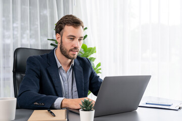Businessman in black suit working on laptop at his workspace desk. Smart executive researching financial data and planning marketing strategy on corporate laptop at modern workplace. Entity