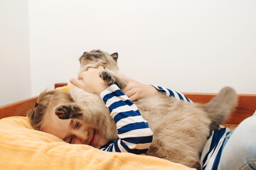 Cute boy plays with a cat at home. Happy kid hugging his cat. Boy relaxing on the bed with pet.