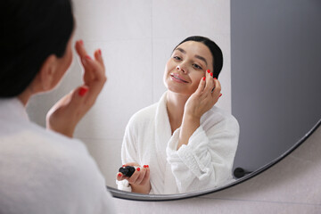 Young woman wearing a bathrobe looking at the round shaped mirror and applying anti-aging skincare to her face. Morning beauty routine concept. Close up, copy space, background.