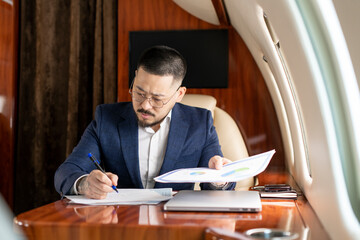 successful Asian businessman in suit and glasses sits in private jet and works with documents,...