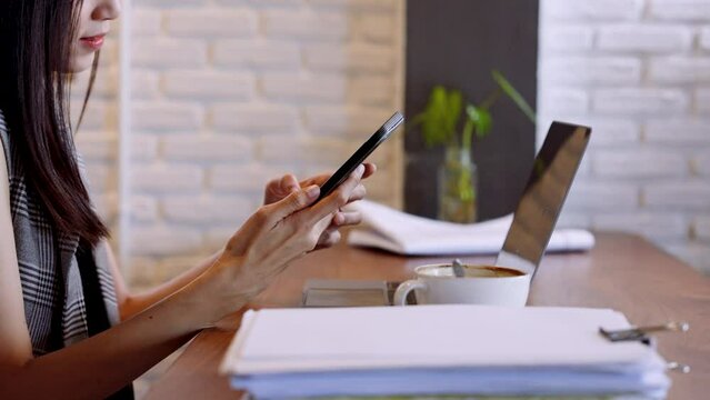 Hand close-up of working day asian woman Using a finger to press a mobile phone to chat with friends while Sitting at work with a laptop computer and there is a stack of documents in bakery shop