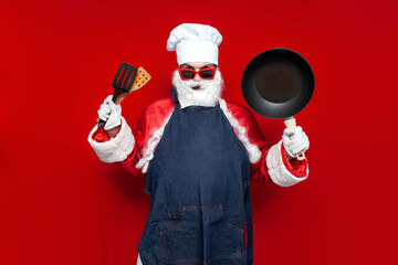 santa claus in chef's hat and apron holds frying pan and utensils on red background, male cook in...