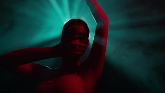 Young dancer performs contemporary dance solo routine. Choreographer gestures hands in modern dance show on dark stage with colorful dramatic lighting. Graceful ballerina dancing in neon light