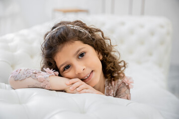 portrait of positive middle eastern girl with brunette curly hair posing in floral dress and leaning on white couch inside of luxurious wedding salon, smiling kid, blurred background