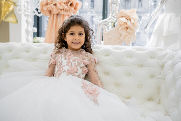 cheerful middle eastern girl with curly hair sitting in floral dress on white couch and smiling inside of luxurious wedding salon, smiling kid, tulle skirt, blurred background