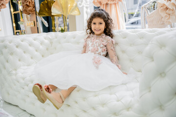 cheerful middle eastern girl with curly hair sitting in floral dress with tulle skirt and shoes on white couch inside of luxurious wedding salon, smiling kid, blurred background