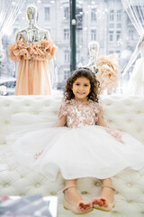 Fototapeta na wymiar cheerful middle eastern girl with curly hair sitting in floral dress on white couch inside of luxurious wedding salon, smiling kid, tulle skirt, bridal, blurred mannequin on background