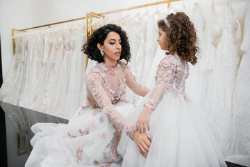special moment, brunette middle eastern bride in floral wedding gown adjusting skirt of her little daughter`s attire in bridal salon around white tulle fabrics, bridal shopping, togetherness