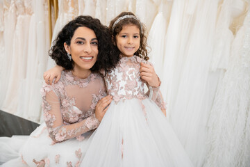 Fototapeta na wymiar special moment, happy middle eastern woman in floral wedding gown sitting and embracing her little daughter in bridal salon around white tulle fabrics, bridal shopping, togetherness, bride-to-be
