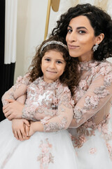 cheerful middle eastern bride in wedding dress hugging happy girl in cute floral attire in bridal salon, shopping, special moment, mother and daughter, happiness, looking at camera