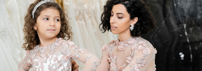 elegant middle eastern bride with brunette wavy hair in wedding dress adjusting cute floral attire of daughter in bridal salon, shopping, special moment, blurred white gown, banner