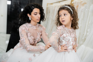 blurred middle eastern bride with brunette wavy hair in wedding dress adjusting cute floral dress of surprised daughter in bridal salon, shopping, special moment, hands on hips, white gown