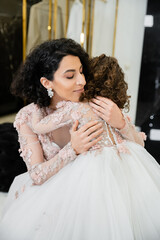 Fototapeta premium charming middle eastern bride with wavy hair in stunning wedding dress hugging daughter in cute floral attire in bridal salon, shopping, happiness, special moment, togetherness, blissful