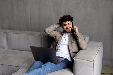 Young handsome man sitting on couch in living room at home using computer chatting and holding smartphone.