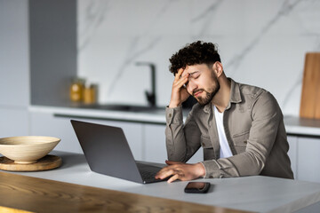 Young man in glasses sit at table looks at laptop screen touch forehead feels confused has business or pc problems