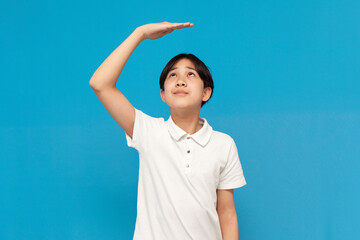 asian boy of twelve years old measures his height with his hand on blue isolated background, korean not tall child - obrazy, fototapety, plakaty