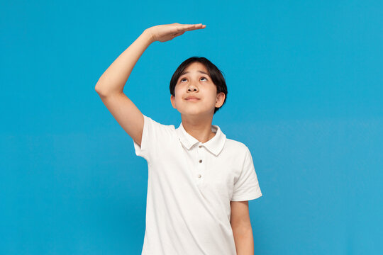 Asian Boy Of Twelve Years Old Measures His Height With His Hand On Blue Isolated Background, Korean Not Tall Child