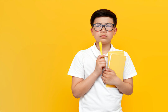 little asian schoolboy holding pen and books and thinking over yellow background, pensive korean kid