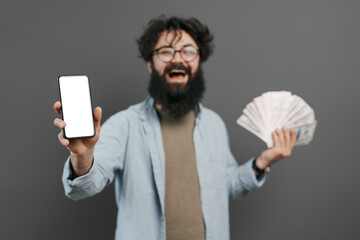 Joyful bearded man in casual clothes posing isolated on grey wall background, studio portrait. People lifestyle concept. Holding mobile phone, fan of cash money in dollar banknotes