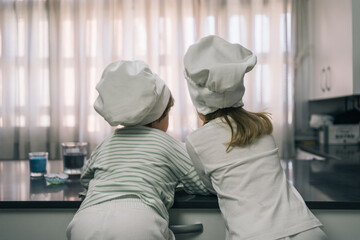 Couple of blond children in chef hats with their backs turned and sitting on their knees on the...