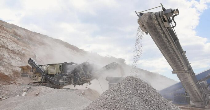 Stone crushing machine at open pit mining and processing plant for crushed stone. Piles of gravel stone and conveyor belts on the side of the road, slow motion