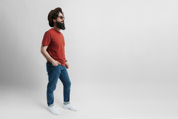 Full length photo of cheerful bearded man on white background. He is wearing an red t-shirt a black hat and stylish sunglasses