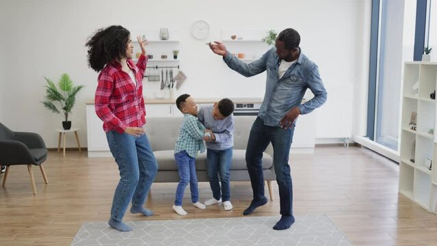 Dynamic multiethnic family of four having dancing party in bright living room of modern apartment. Healthy young parents wearing jeans teaching sons different styles of movements at leisure time.