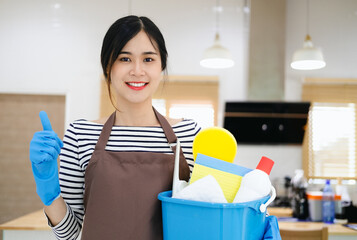 Smiling young woman housewife in brown apron and blue gloves holding cleanser bottles and showing thumb up , Standing in modern kitchen, professional cleaning service.