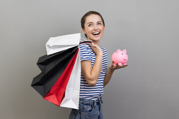 Portrait of excited positive woman wearing striped T-shirt holding shopping bags and piggy bank,...