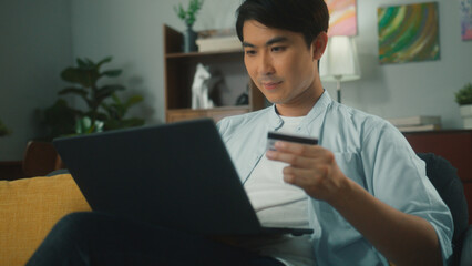 Attractive young asian man shopping online using laptop computer holding credit card purchase via e-banking. Handsome guy making online payment through e-commerce platform on sofa in cozy living room.
