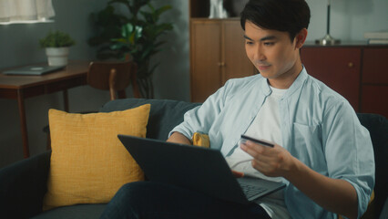Attractive young asian man shopping online using laptop computer holding credit card purchase via...