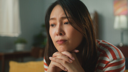 Portrait of thoughtful attractive young Asian woman props head with hand feeling bored and sad. Exhausted beautiful female have nothing to do with absent minded sit on couch at cozy living room
