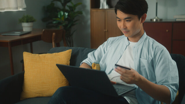 Attractive Young Asian Man Shopping Online Using Laptop Computer Holding Credit Card Purchase Via E-banking. Handsome Guy Making Online Payment Through E-commerce Platform On Sofa In Cozy Living Room.