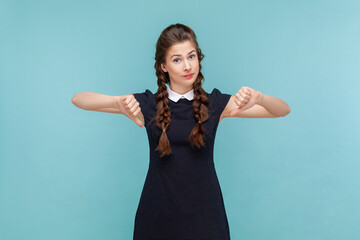Portrait of displeased naughty woman with braids standing and showing thumb down, dislike bad service, not recommend, wearing black dress. woman Indoor studio shot isolated on blue background.
