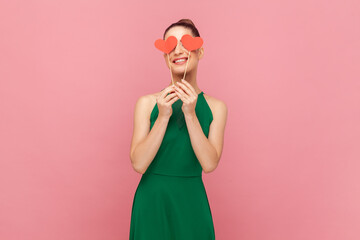 Portrait of charming unknown woman with bun hairstyle standing and covering her eyes with little red hearts on stick, smiling, wearing green dress. Indoor studio shot isolated on pink background.