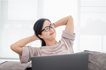 Happy relaxed mature woman sitting in her kitchen with a laptop in front of her stretching her arms above her head and looking out of the window with a smile