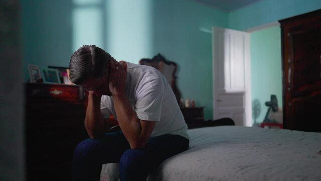 Depressed man sitting by bedside in bedroom. Sad unhappy person looking down suffering from mental illness