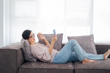 Pleasant senior mature lady resting on couch, using applications on smartphone. Happy old mature woman chatting in messenger or social network with friends or children, shopping in internet store.