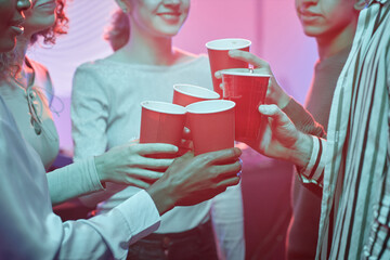 Closeup of young people at house party toasting with red cups in neon lights, copy space