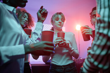 Diverse group of young people at house party holding red cups with alcohol and dancing in neon light