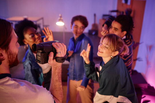 Group of young people dancing at house party with man holding retro video camera and filming home video of friends having fun, copy space